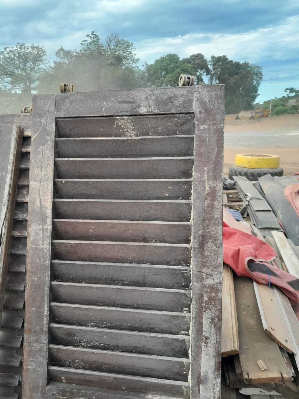 Painel metálico gradeado (portão/persiana industrial) usado disponível no pátio, entre outras peças de madeira e materiais reaproveitados de obra.