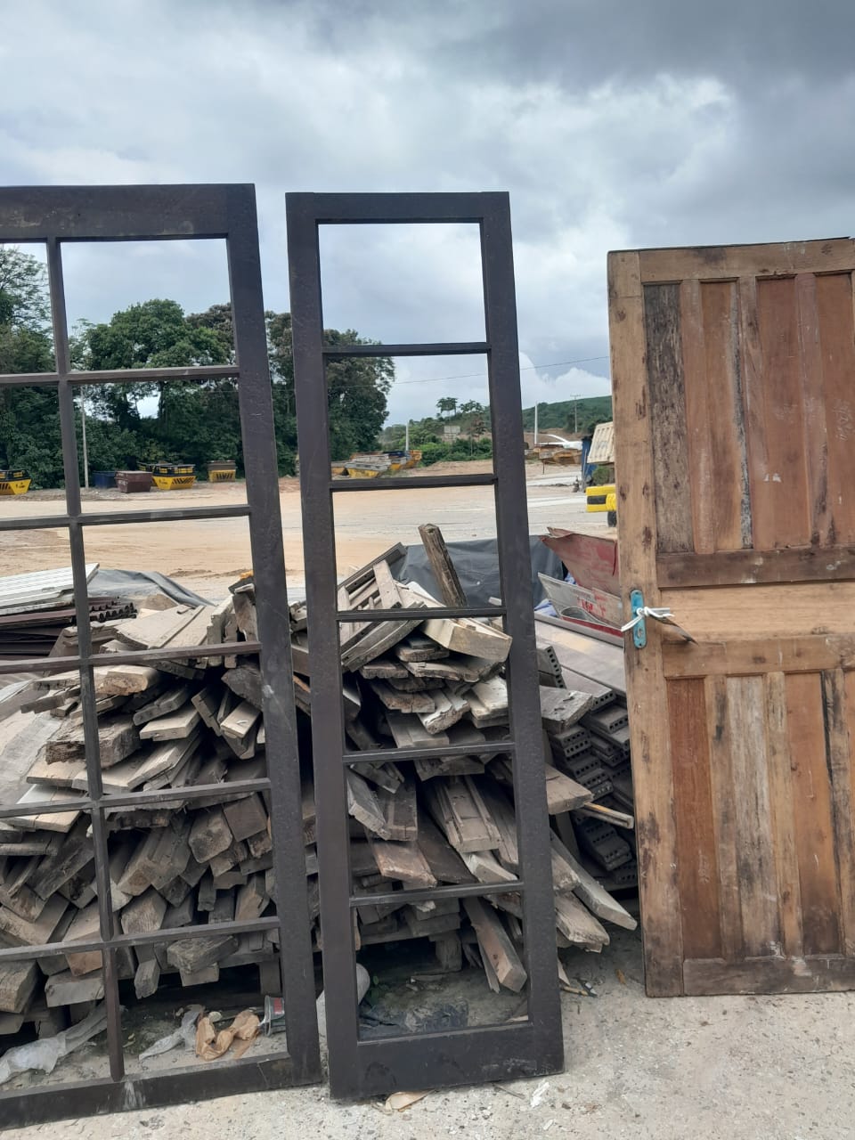 Pátio com estoque de materiais reaproveitados: esquadrias metálicas, porta de madeira maciça e pilhas de pranchas/tábuas de demolição à venda.