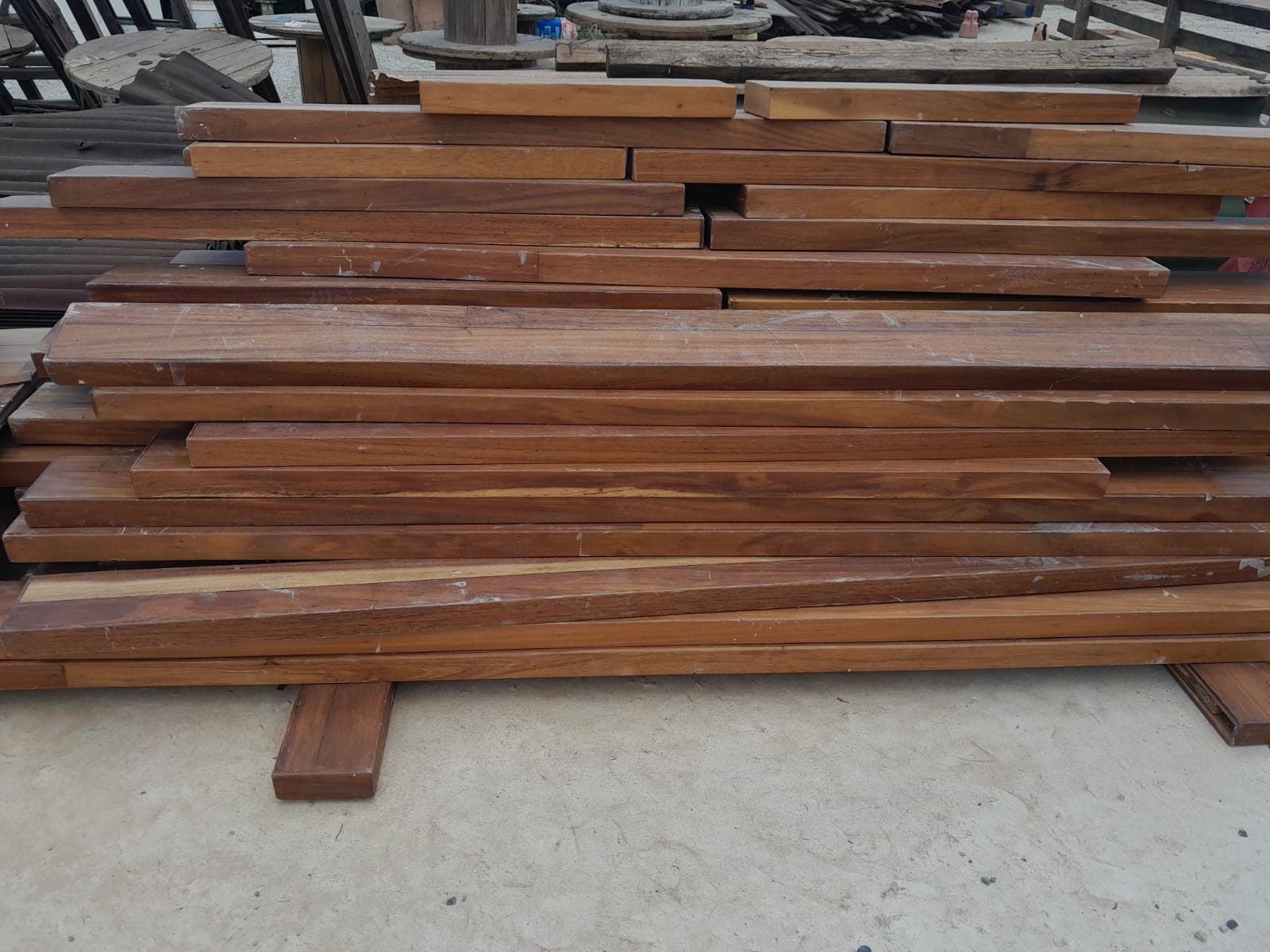 Estoque de tábuas e pranchas de madeira de demolição (hardwood) empilhadas, com carretéis reaproveitados ao fundo, mostrando o material disponível para venda.