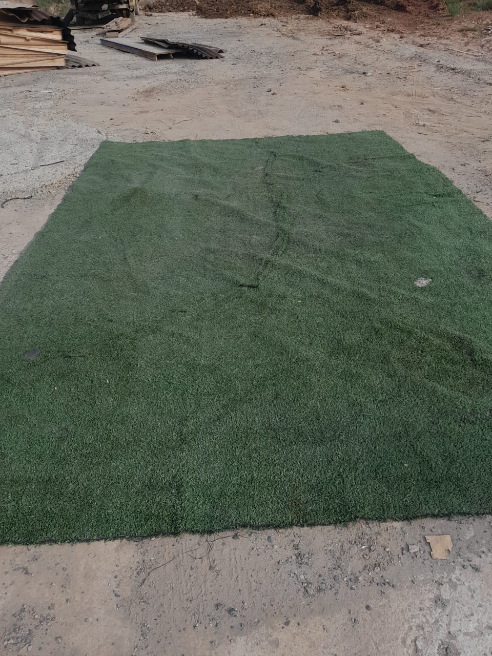 Tapete de grama sintética verde disponível para venda no pátio, com estoque de madeiras reutilizadas ao fundo.